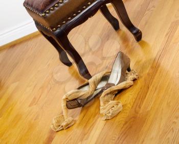 Angled photo of woman stockings and dress shoes lying next to padded footstool with red oak floors in background 