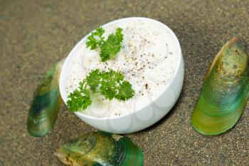 Closeup angled horizontal photo of fresh clam chowder in a white cup plus parsley and pepper with calm shells and sand surrounding outside of bowl   