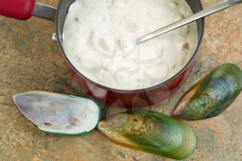 Closeup horizontal photo of clam chowder in sauce pan with clam shells on side all on top of a natural stone counter top