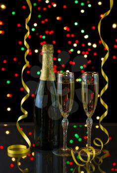 Vertical photo of a bottle of champagne, two tall elegant glasses with yellow ribbons and lights in background