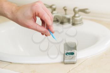 Horizontal photo of male hand picking up dental tooth pick in bathroom with dental floss container, sink and counter top in background 