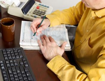 Horizontal photo of mature man, partial face view, working on his taxes with calculator, tax tables and other office equipment in background 