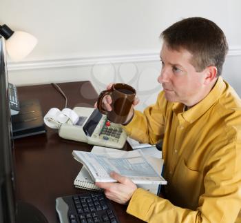 Photo of mature man, looking at data on computer monitor, working on his taxes while holding cup of coffee with office equipment in background 