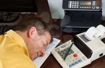 Horizontal photo of mature man relaxing, head position on tax forms, while working on his taxes with calculator with other office equipment in background 