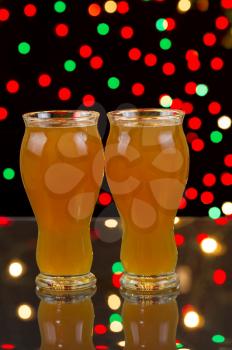 Vertical photo of two tall glasses, on glass table, filled with premium Amber Ale with party lights in background 