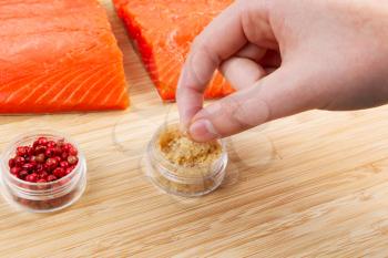 Horizontal photo of female fingers pinching sea salt, from container, with fresh pieces of Wild Red Salmon and natural bamboo board in background 