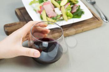 Closeup photo of female hand holding red wine glass with freshly made salad on white plate with fork in background 