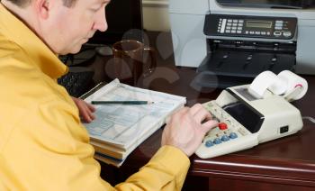 Horizontal photo with focus on male hand while using calculator with tax tables and other office equipment in background 
