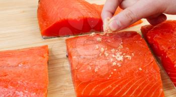 Horizontal photo of female fingers placing sea salt on fresh pieces of Wild Red Salmon and natural bamboo board in background 