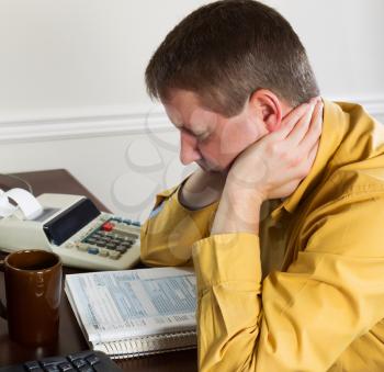 Photo of mature man, holding head with hands, working on his taxes with tax booklet and office equipment in background 