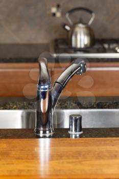 Vertical photo of a chrome kitchen faucet with stone counter tops, cherry siding and range in background  