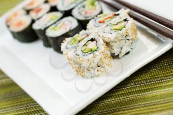 Closeup horizontal angled photo of freshly handmade California sushi rolls, filling white plate, and chop sticks in background with texture green cloth underneath 