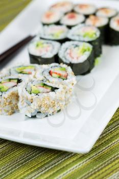 Closeup vertical photo of freshly handmade sushi rolls, filling white plate, and chop sticks in background with texture green cloth underneath 