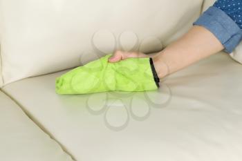 Horizontal photo of female hand holding microfiber rag with cleaning solution in front of white leather sofa 