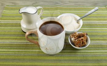 Closeup horizontal photo of a full cup of coffee with sugar and spoon in bowl, snacks and cream in pouring spout with green striped textured cloth underneath 