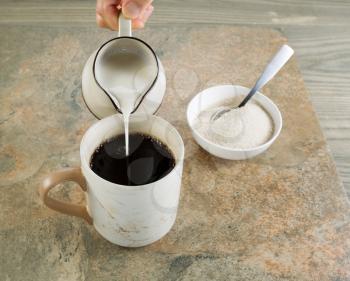 Photo of female hand pouring cream into a cup of black coffee with stone and aged wood underneath