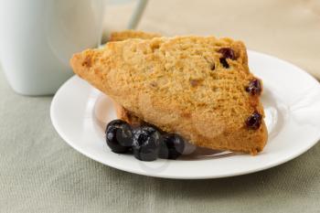Closeup horizontal photo of scones on white plate with blue berries up front and tea cup plus napkin in background