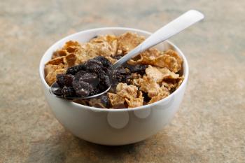 Horizontal photo of grain cereal, raisins, spoon in white bowl with stone counter top understand