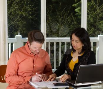 Photo of mature woman showing man where to sign document while working at home with laptop, calculator and papers on top of table and large windows in background