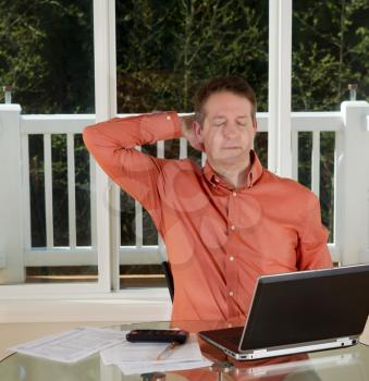 Photo of mature man resting while working from home with laptop, calculator and papers on top of table and large windows in background

