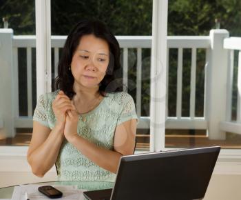 Photo of mature woman looking at computer screen while working at home with laptop, cell phone and papers on top of table and large windows in background