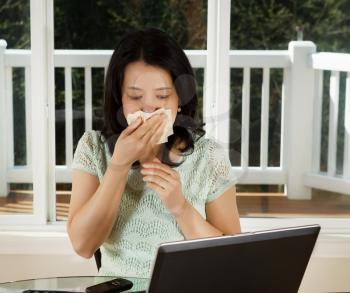 Photo of mature woman wiping her nose with tissue while working at home with laptop and cell phone on top of table and large windows in background