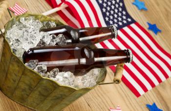 Angled horizontal photo of glass bottled beer in old metal bucket filled with ice and American flags in background for celebration of Independence Day