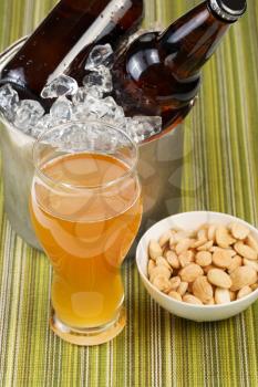 Closeup vertical photo of a golden beer in tall glass with salted nuts in small bowl with bottled beer in ice bucket in background