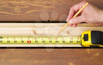 Horizontal photo of hand holding pencil next to tape measure on top of new cedar wood board next to fading wood on outdoor wooden deck 
