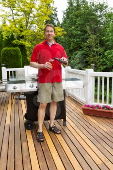Closeup vertical photo of mature man pouring beer into glass with BBQ grill and open cedar patio with seasonal trees in full bloom in background 