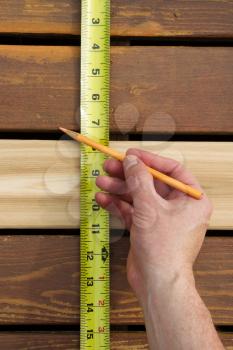 Vertical photo of hand with pencil and tape measuring space between new cedar wood boards on outdoor wooden deck 
