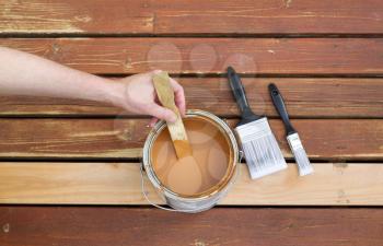 Horizontal photo of male hand stirring wood stain in can with stir stick with two paint brushes lying on a single new cedar wooden board next to fading wood on outdoor deck 