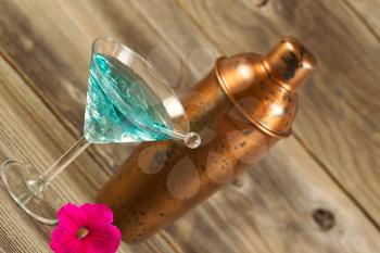 Angled horizontal view of a mixed drink, bright pink flower, stir stick and a metal mixer resting on rustic wood