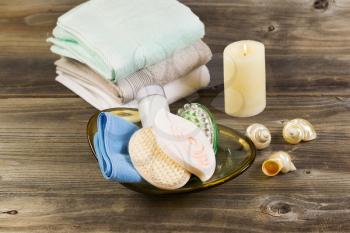 Horizontal front top view of spa hygiene accessories in glass bowl, light white candle with clean stacked towels in background and seashells on rustic wood