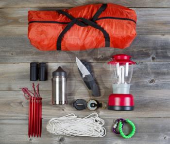 Overhead view of basic hiking gear placed on weathered wooden boards. Items include tent inside of bag, pegs, compass, canteen, rope, knife, case, lantern and binoculars. 