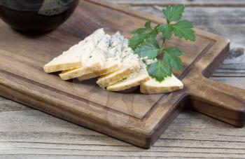 Closeup horizontal photo of sliced blue cheese, parsley on the side, on a traditional wooden server with a partial glass of red wine 