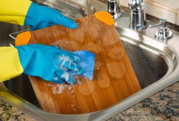 Closeup horizontal image of hands wearing rubber gloves washing bamboo cutting board with sponge and soapy water inside of kitchen sink 