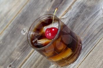 Closeup horizontal angled view of a rum and cola with large red cherry and ice in glass position on rustic wood 