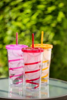 Vertical photo of colorful plastic cups filled with ice and water on table with green blurred out trees in background 