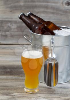 Vertical view of a freshly pour gold beer with bottled beer sitting inside metal bucket filled with crushed iced and rustic wood in background 