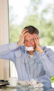 Vertical image of mature man showing stress by biting wad of paper while working from home with bright daylight coming in from window in background