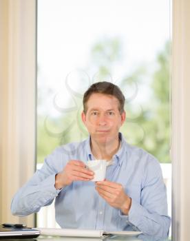 Vertical image of mature man looking forward while holding a cup of coffee with bright daylight coming in from window in background