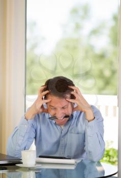 Vertical image of mature man showing extreme stress by biting his pen while working from home with bright daylight coming in from window in background
