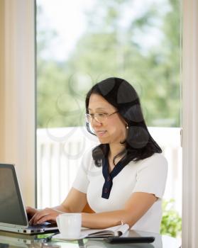 Vertical image of mature woman relaxed during typing on laptop while working from home with blurred out daylight coming in from window in background