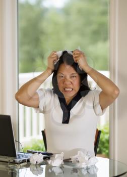 Vertical image of mature woman angrily putting ripped paper up to her forehead while working from home with blurred out daylight coming in from window in background