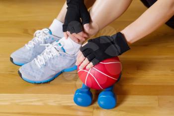 Horizontal image of female hands wearing workout gloves while resting hand on small weight ball with wooden gym floor and partial body in background 