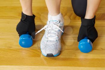 Horizontal image of female hands picking up small dumbbell weights while squatting on wooden gym floor 
