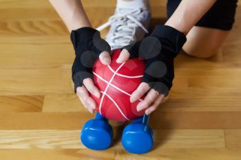 Horizontal image of female hands wearing workout gloves while lifting red weight ball of off blue dumbbells on wooden gym floor 