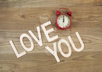Overhead view of an old table top alarm clock and large wooden letters spelling out Love You on rustic wood