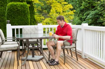 Horizontal photo of mature man holding a glass of orange juice while preparing to read a magazine on outdoor patio with green and yellow trees in full seasonal bloom 